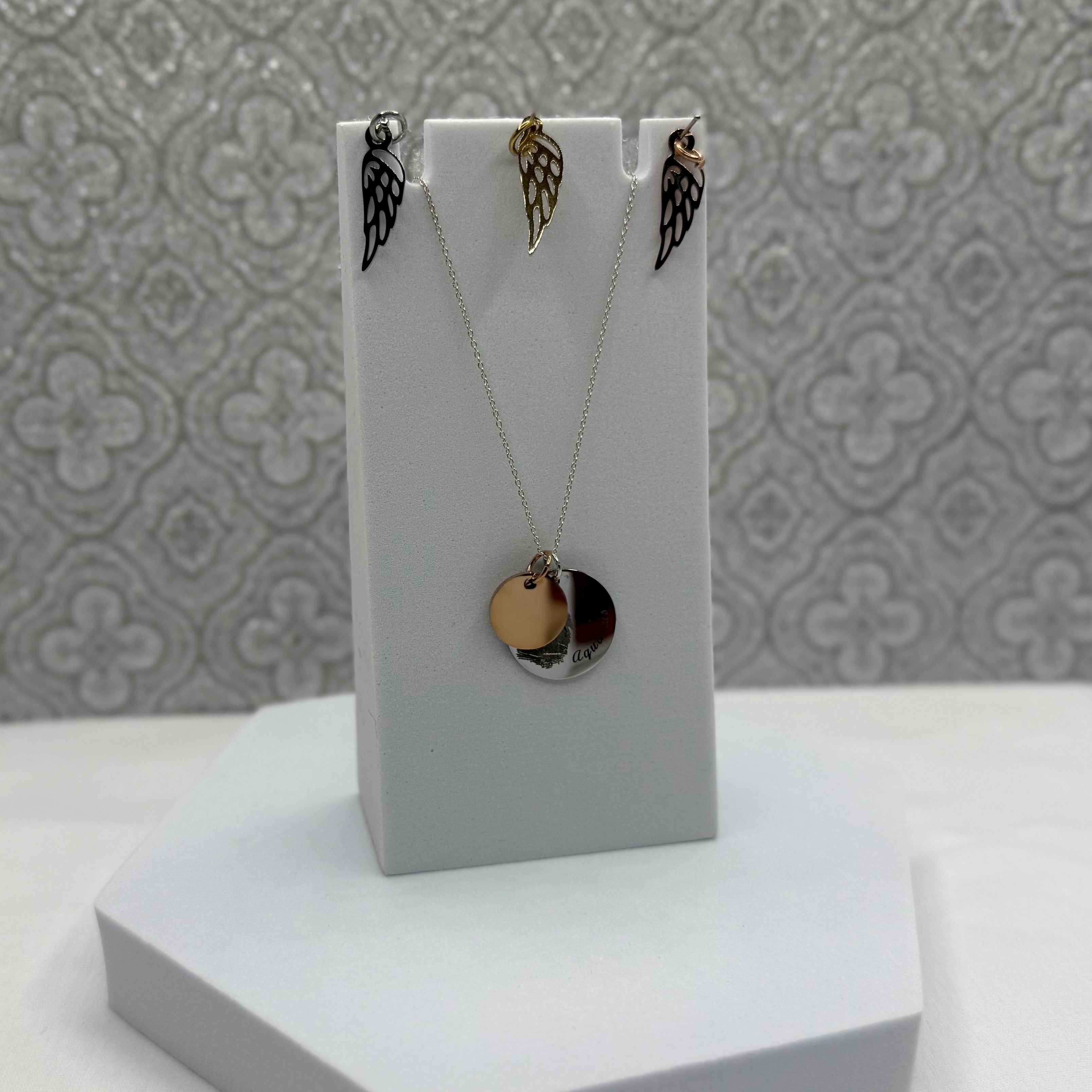 19mm Disc Pendant in Stainless Steel with a fingerprint and Aquarius curved along the edge. It is paired with a 13mm Rose Gold Coated Stainless Steel Disc that can also be engraved with Initials or a small design of your choice. Above the necklace is 3 Angel Wing Charms we offer in Stainless Steel, Gold Coated Stainless Steel, and Rose Gold Coated Stainless Steel.