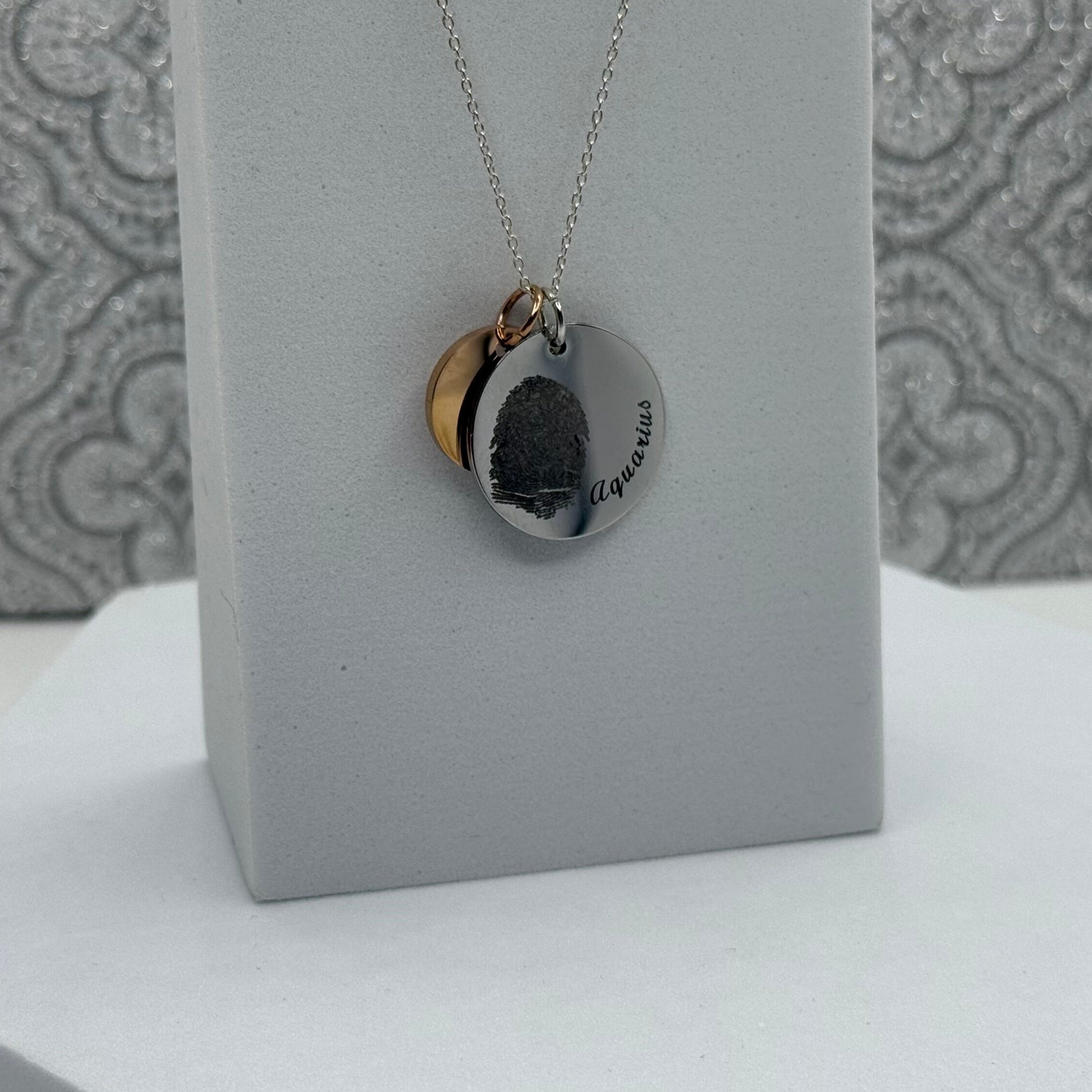19mm Disc Pendant in Stainless Steel with a fingerprint and Aquarius curved along the edge. It is paired with a 13mm Rose Gold Coated Stainless Steel Disc that can also be engraved with Initials or a small design of your choice.